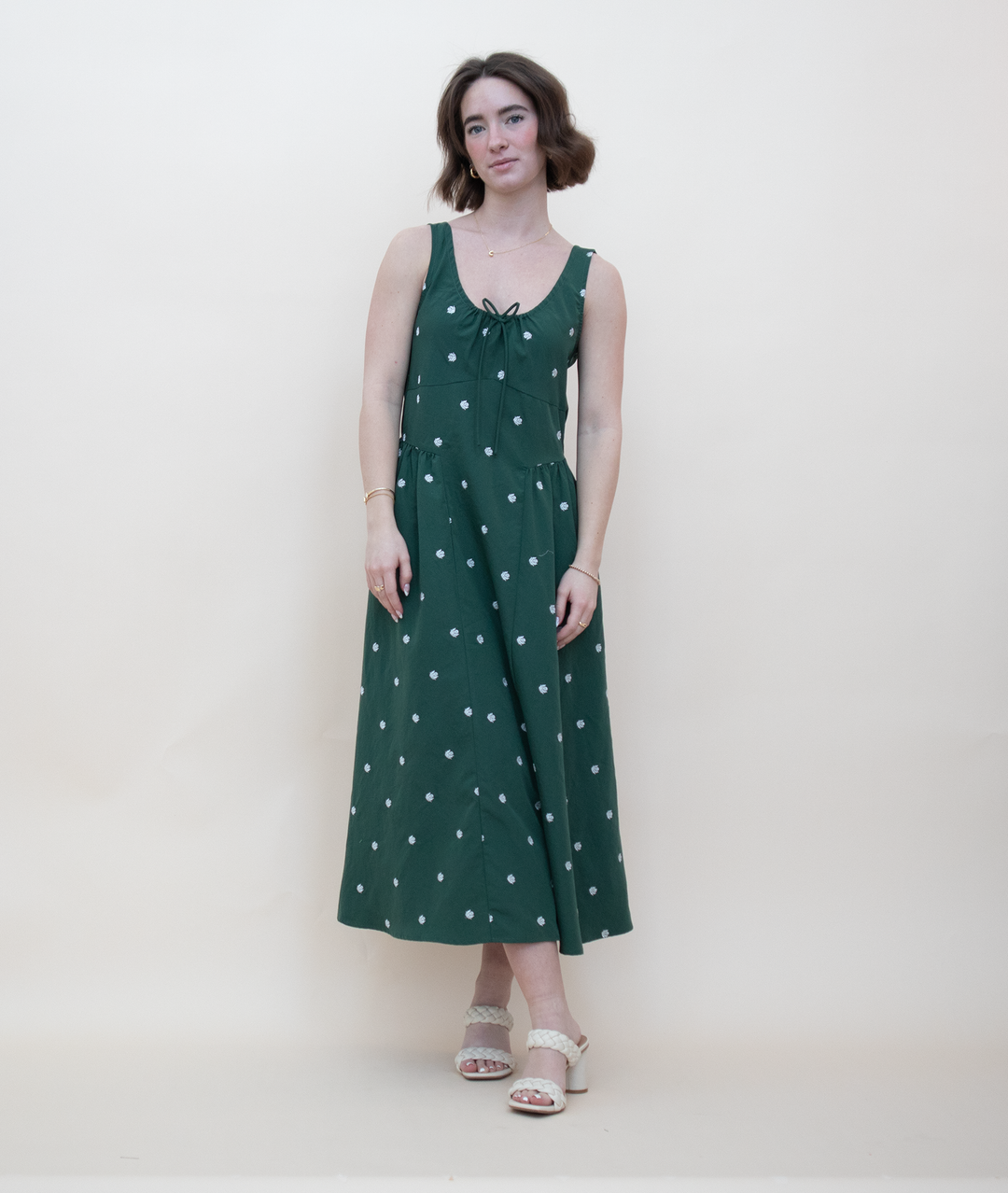 Women's dark green sleeveless midi dress with white daisy floral print, gathered tie neckline, and A-line silhouette, styled with cream block heel sandals