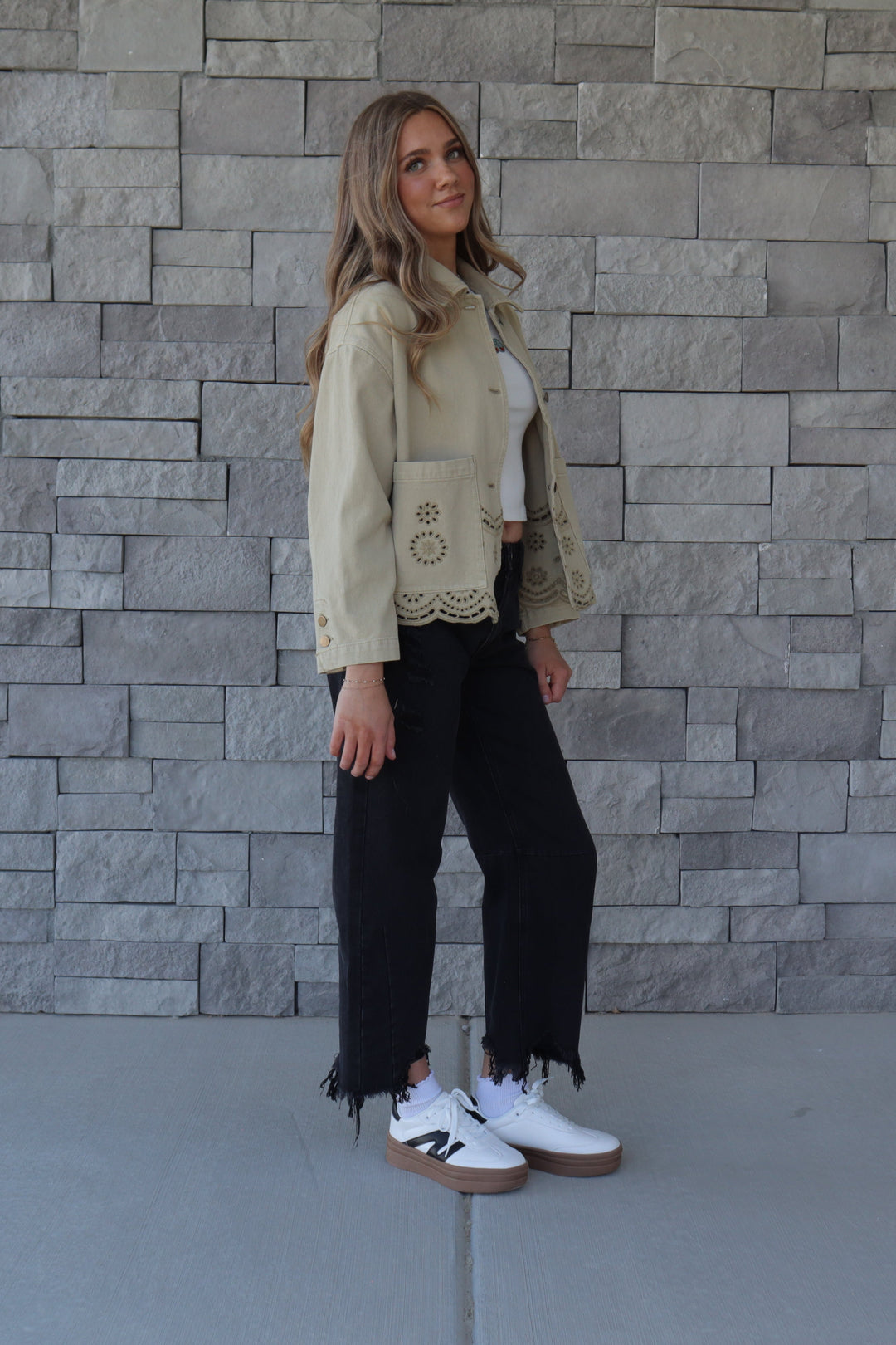 A woman stands outdoors against a gray stone wall wearing a olive green cropped jacket with lace trim, black cropped pants with fringe, a white top, and white sneakers.