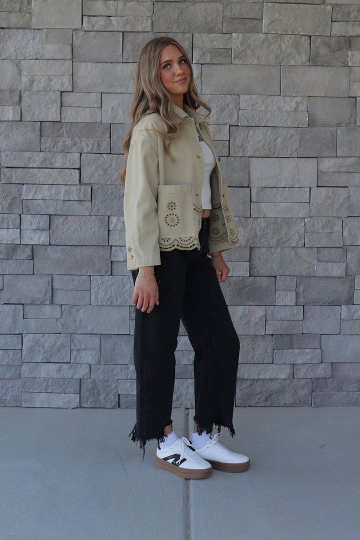 A woman stands outdoors against a gray stone wall wearing a olive green cropped jacket with lace trim, black cropped pants with fringe, a white top, and white sneakers.