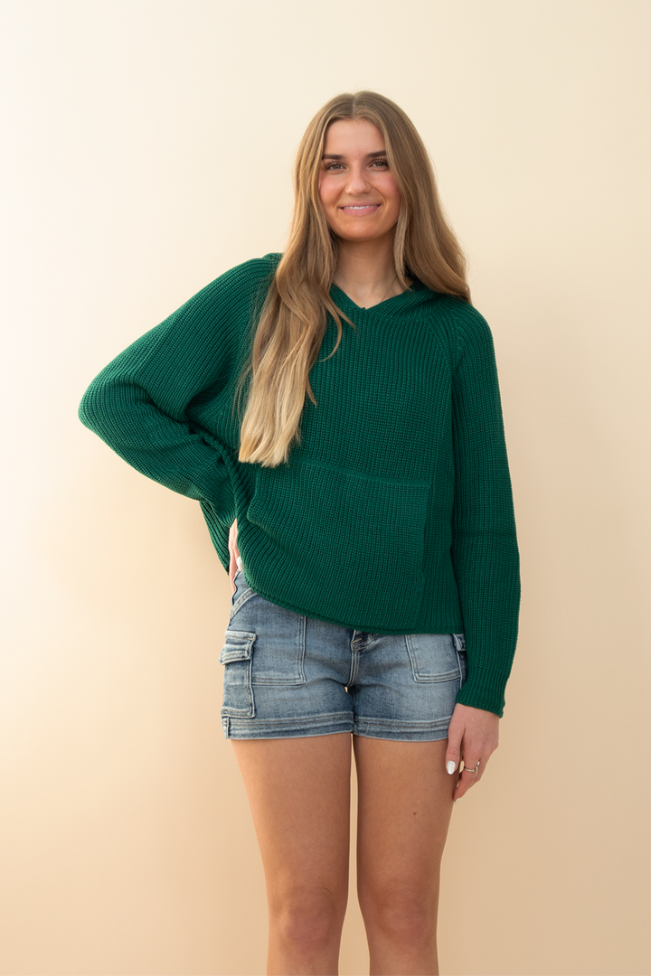 Young woman facing forward in a forest green waffle knit hoodie with front pocket and relaxed hood, paired with light-wash denim cargo shorts and neutral sneakers. Comfortable and casual everyday outfit perfect for transitional seasons, back-to-school fashion, or weekend streetwear looks for Gen Z women.