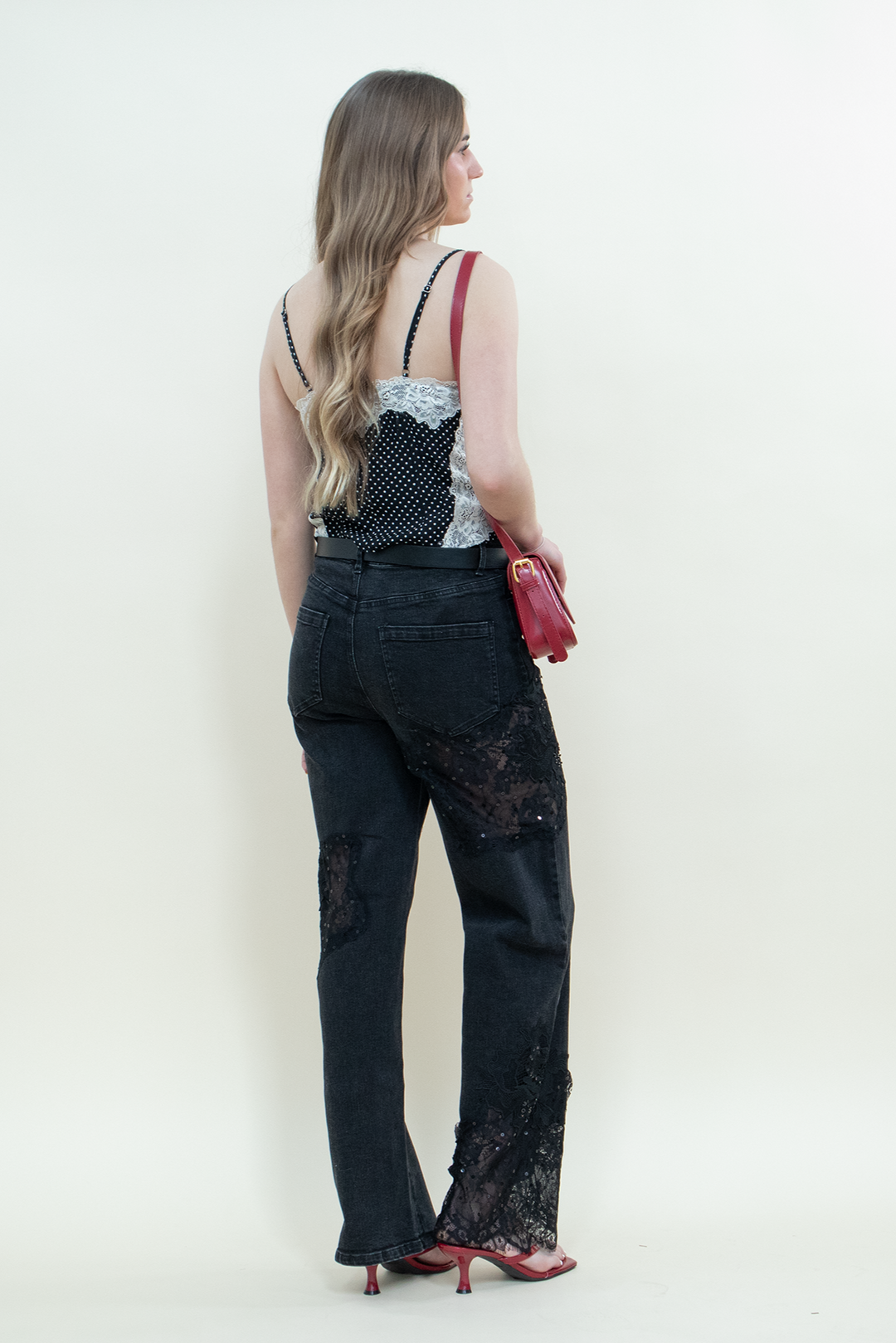 Model facing away in black straight-leg jeans with sheer lace panels from Groove Closet, paired with a black polka dot camisole with white lace trim, red handbag, and red kitten heels.