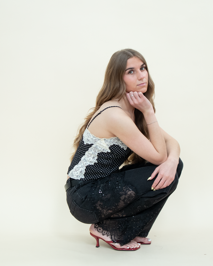 Model squatting in black straight-leg jeans with sheer floral lace panels from Groove Closet, styled with a black polka dot camisole with white lace trim and red kitten heels.