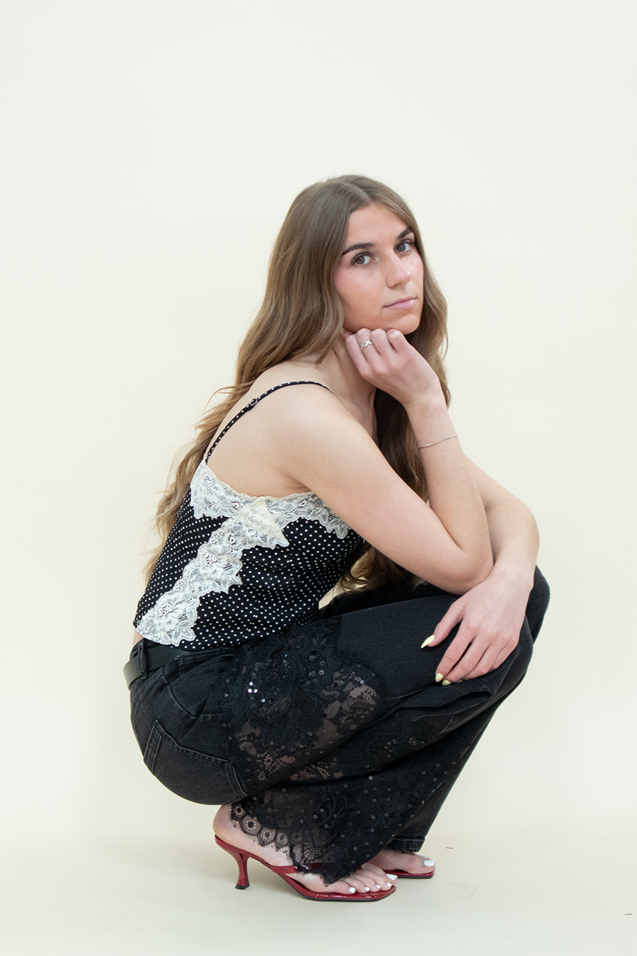Model squatting in black straight-leg jeans with sheer floral lace panels from Groove Closet, styled with a black polka dot camisole with white lace trim and red kitten heels.