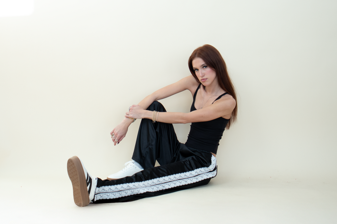 Model seated on the floor wearing relaxed black satin pants with lace stripe down the sides, styled with a fitted black tank top and white sneakers.