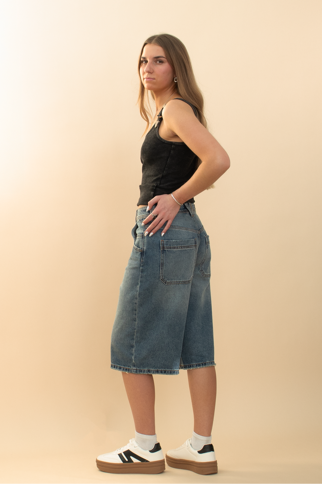 long oversized denim short as seen from the back to show the back pockets, styled with a white sneaker and black tank