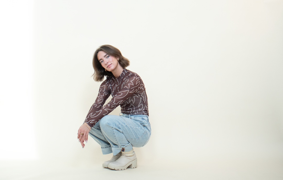 Angled view of a brown sheer mesh top with long sleeves, mock neckline, and cream Western-inspired abstract pattern.