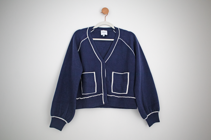 blue cardigan with bishop sleeve and white line detailing hanging from a hanger on a hook.