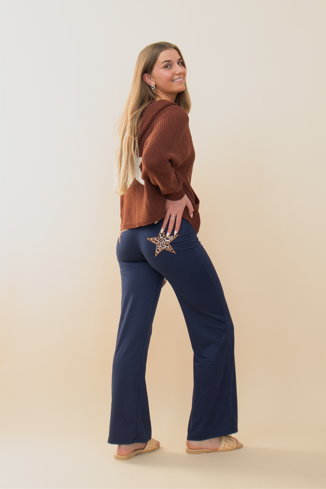 Smiling woman in rust-colored knit sweater and navy blue flare pants with a leopard print star patch on the back pocket, standing against a neutral background.