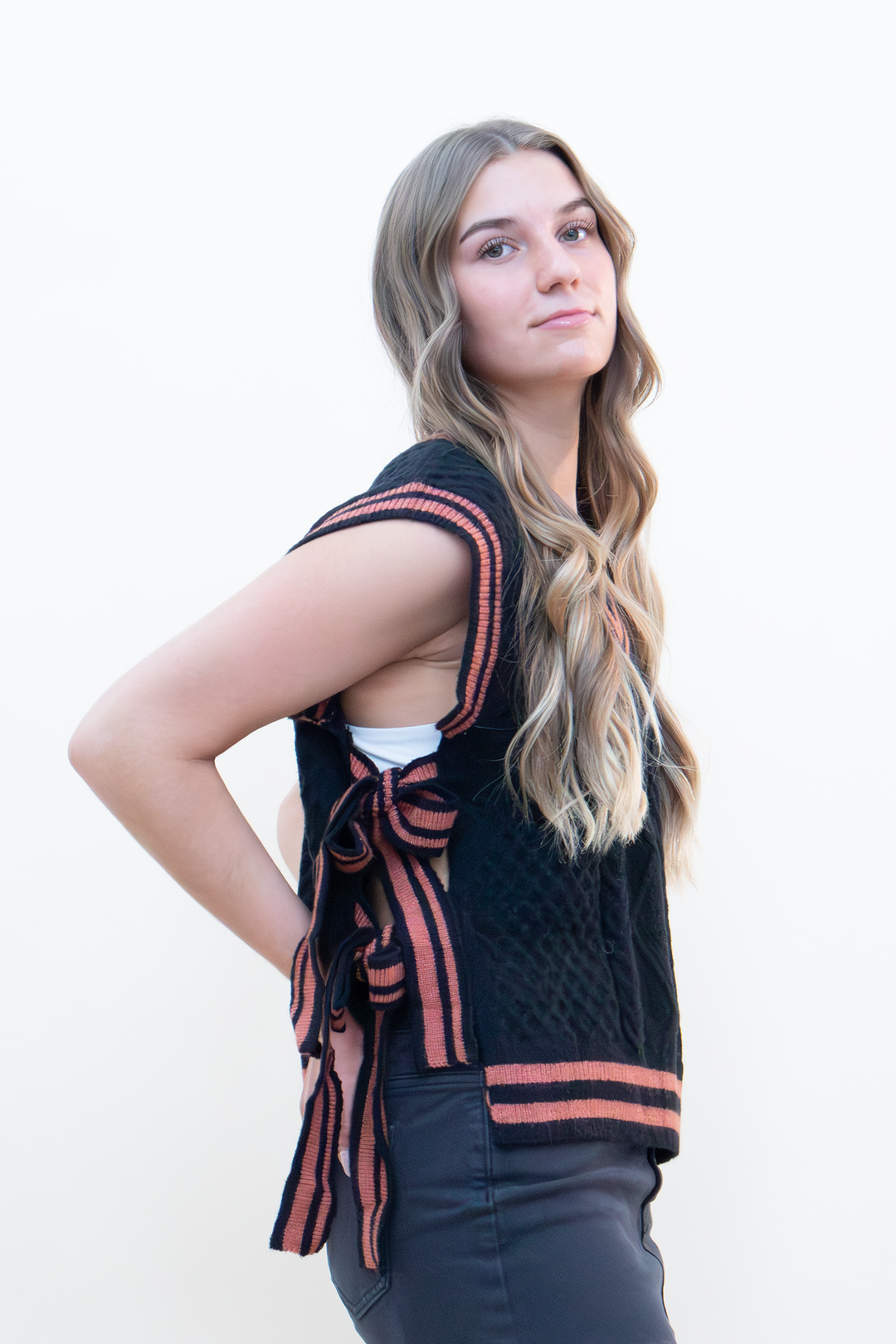 Side view of black cable knit sweater vest with open back and terracotta striped bow tie closure, paired with dark jeans