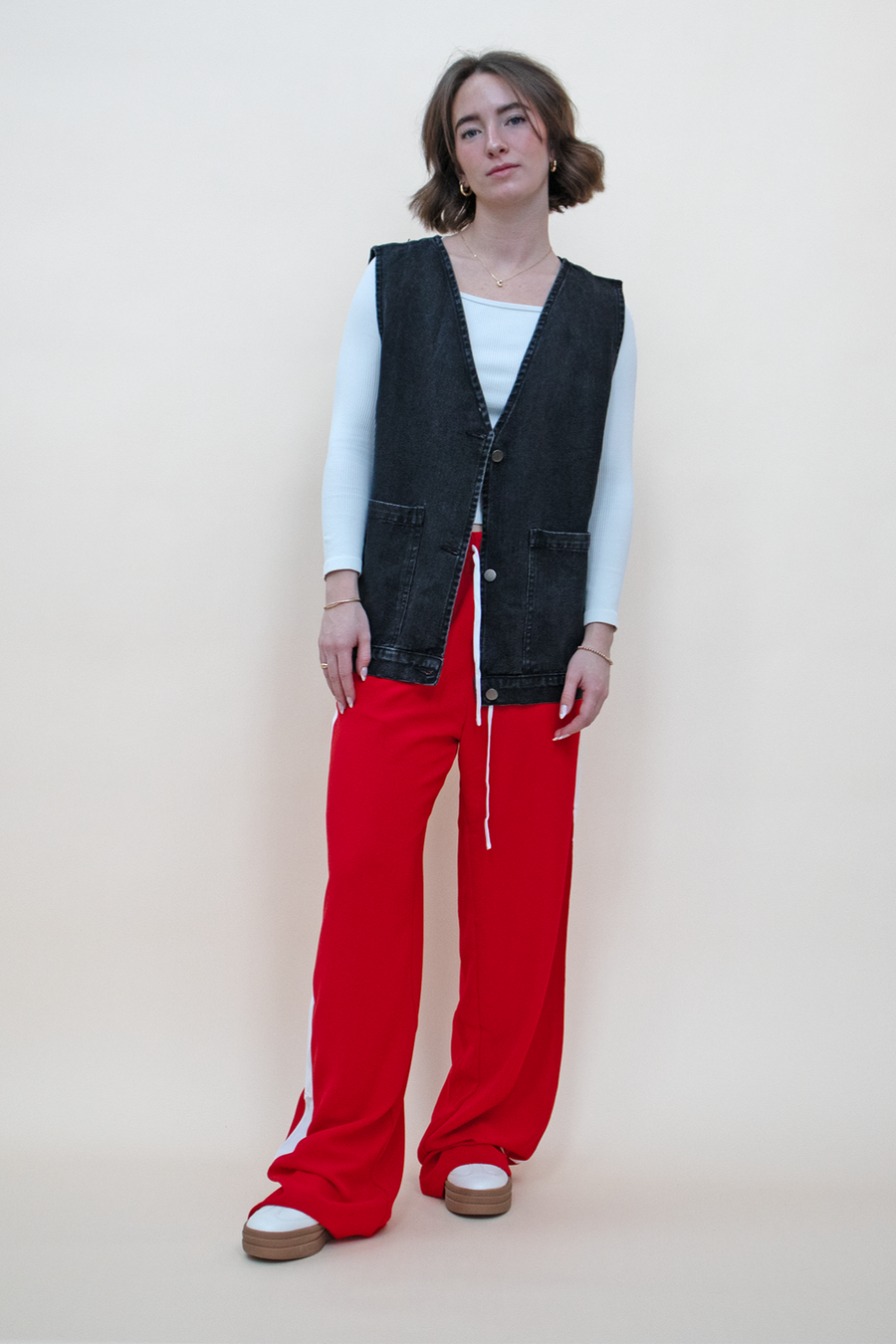 Oversized black denim vest with big pockets and silver buttons, styled with red track pants and a white long sleeve shirt.
