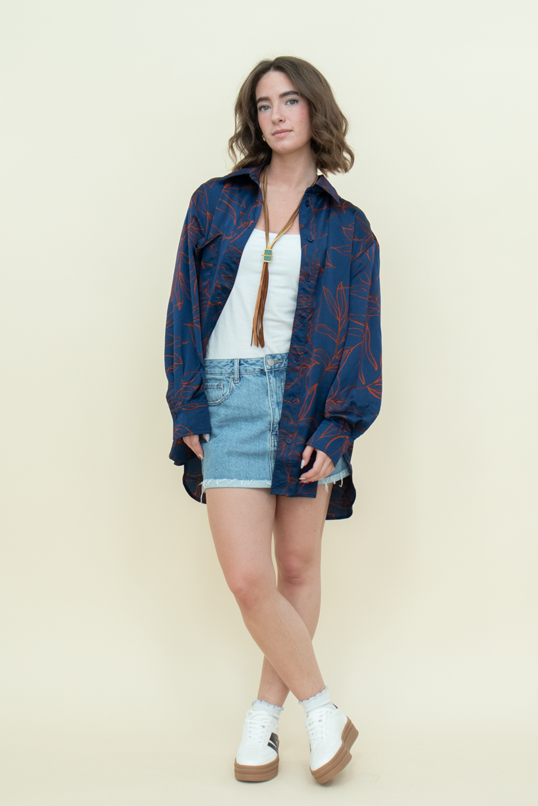 Model wearing a flowy navy leaf-print button-up shirt over a white tank with a light-wash denim mini skirt and white sneakers. Trendy, relaxed streetwear style for women from Groove Closet.