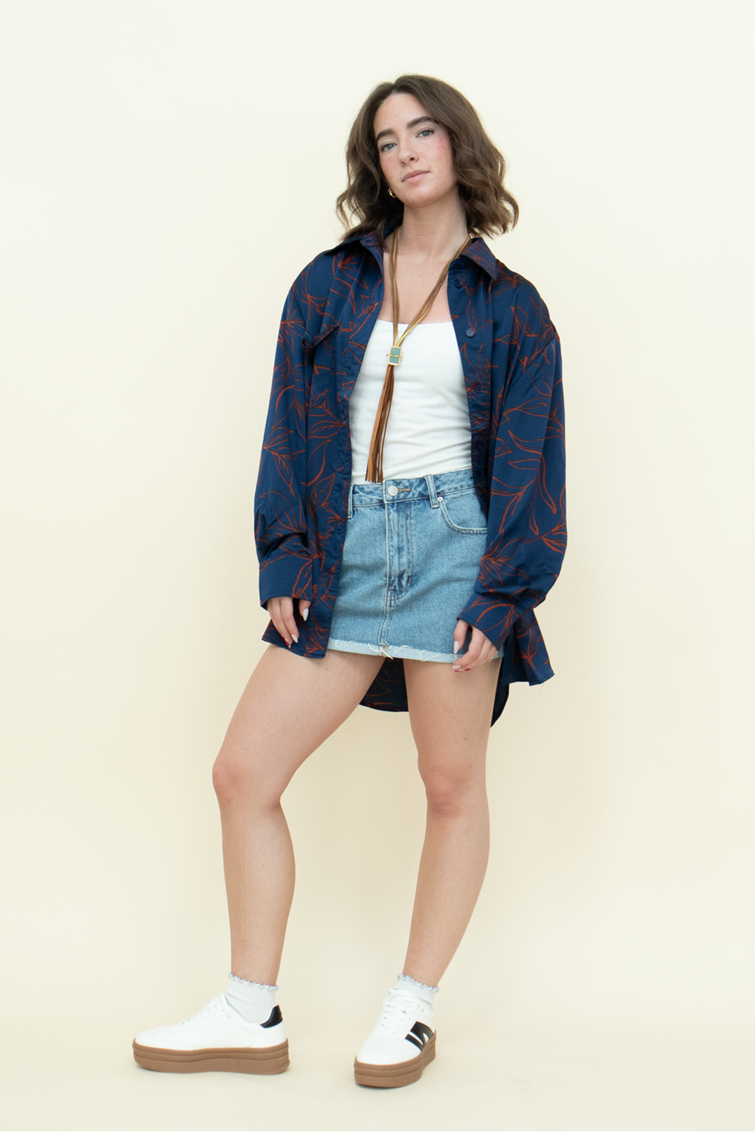Woman in an oversized navy button-up shirt with red leaf print, white tank top, and denim mini skirt styled with white platform sneakers. Casual cool-girl outfit perfect for effortless streetwear looks from Groove Closet.