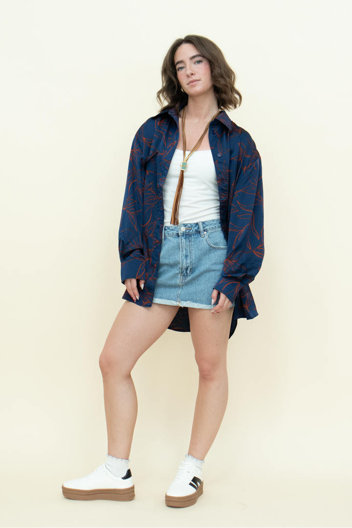 Woman in an oversized navy button-up shirt with red leaf print, white tank top, and denim mini skirt styled with white platform sneakers. Casual cool-girl outfit perfect for effortless streetwear looks from Groove Closet.