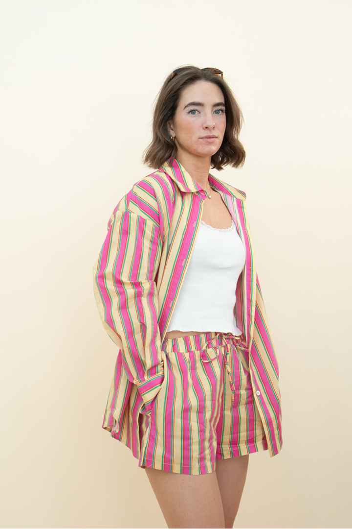 Model wearing Groove Closet’s colorful striped co-ord set featuring a relaxed button-down shirt and matching drawstring shorts, styled with a white tank top and yellow heels – playful summer outfit for women.