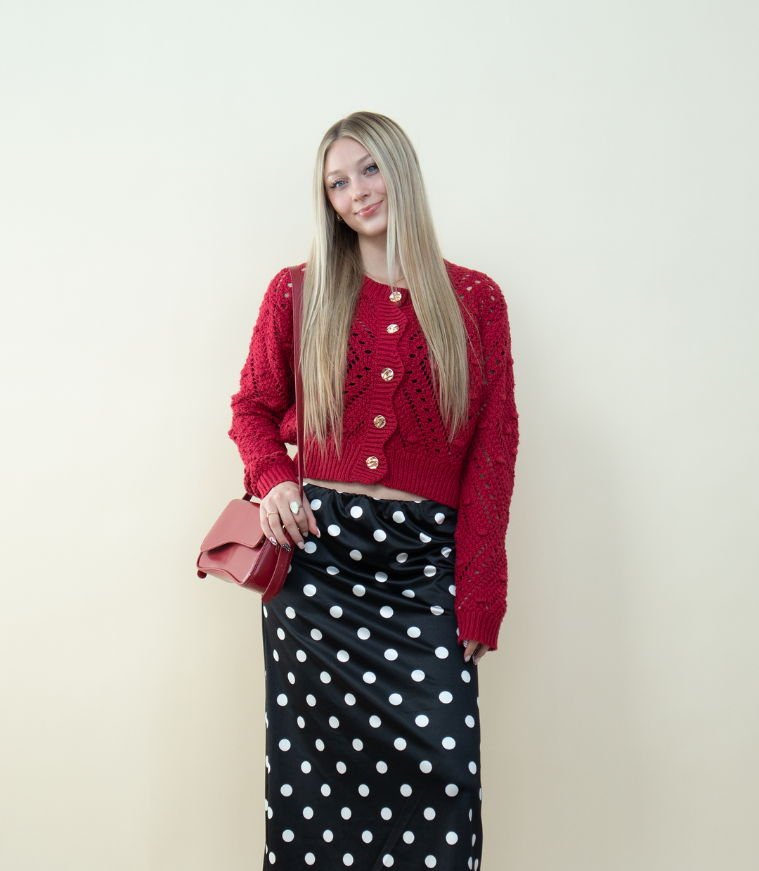 A blonde woman wearing a chunky red knit cardigan with gold buttons styled with a black-and-white polka dot satin skirt and a red crossbody bag. Cozy winter outfit idea featuring a textured red cardigan for trendy feminine fashion.