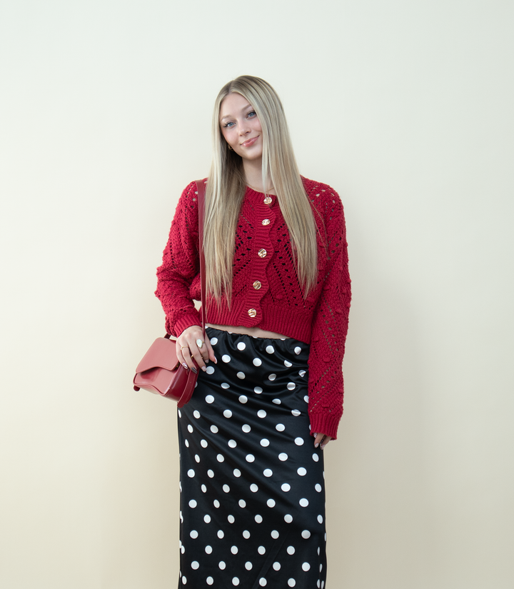 A blonde woman wearing a chunky red knit cardigan with gold buttons styled with a black-and-white polka dot satin skirt and a red crossbody bag. Cozy winter outfit idea featuring a textured red cardigan for trendy feminine fashion.
