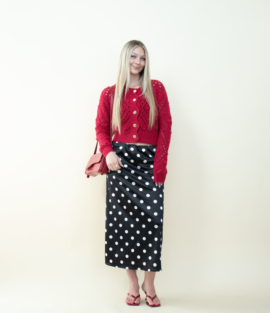 Full-body outfit featuring a red eyelet knit cardigan with statement gold buttons paired with a black polka dot midi skirt and red heels. Classic and chic styling inspiration for a bold red cardigan.