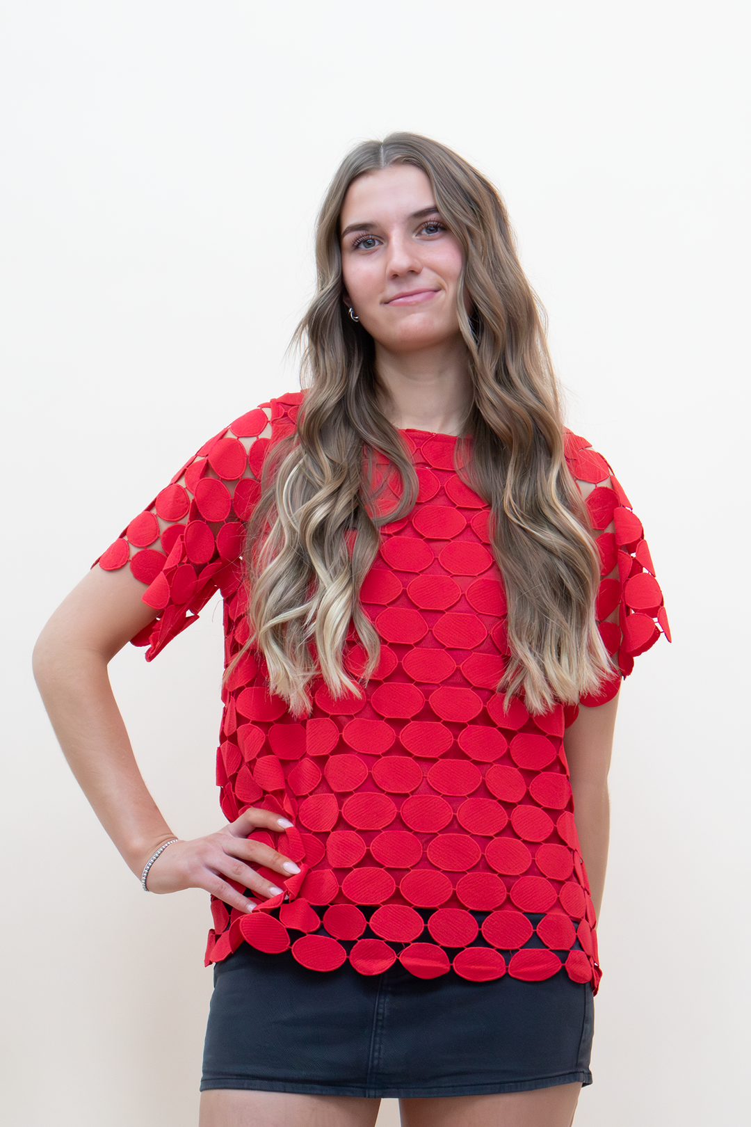 Red oversized short-sleeve top covered in layered oval appliqué patches creating a scale-like texture, crew neckline, cropped front view on model styled with a dark denim mini skirt