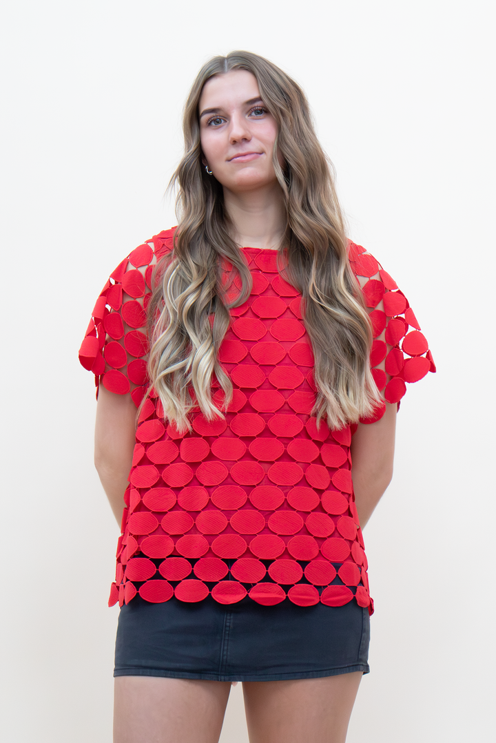 Front view of red short-sleeve boxy top with allover circle appliqué cutwork detailing and scalloped hem, paired with a charcoal denim mini skirt
