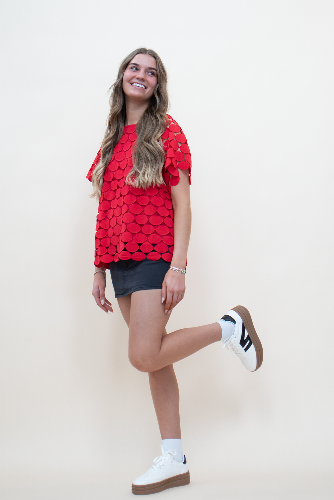 Full-length view of red oval appliqué boxy top with open cutwork construction, styled with a dark denim mini skirt and white platform sneakers, model in playful pose