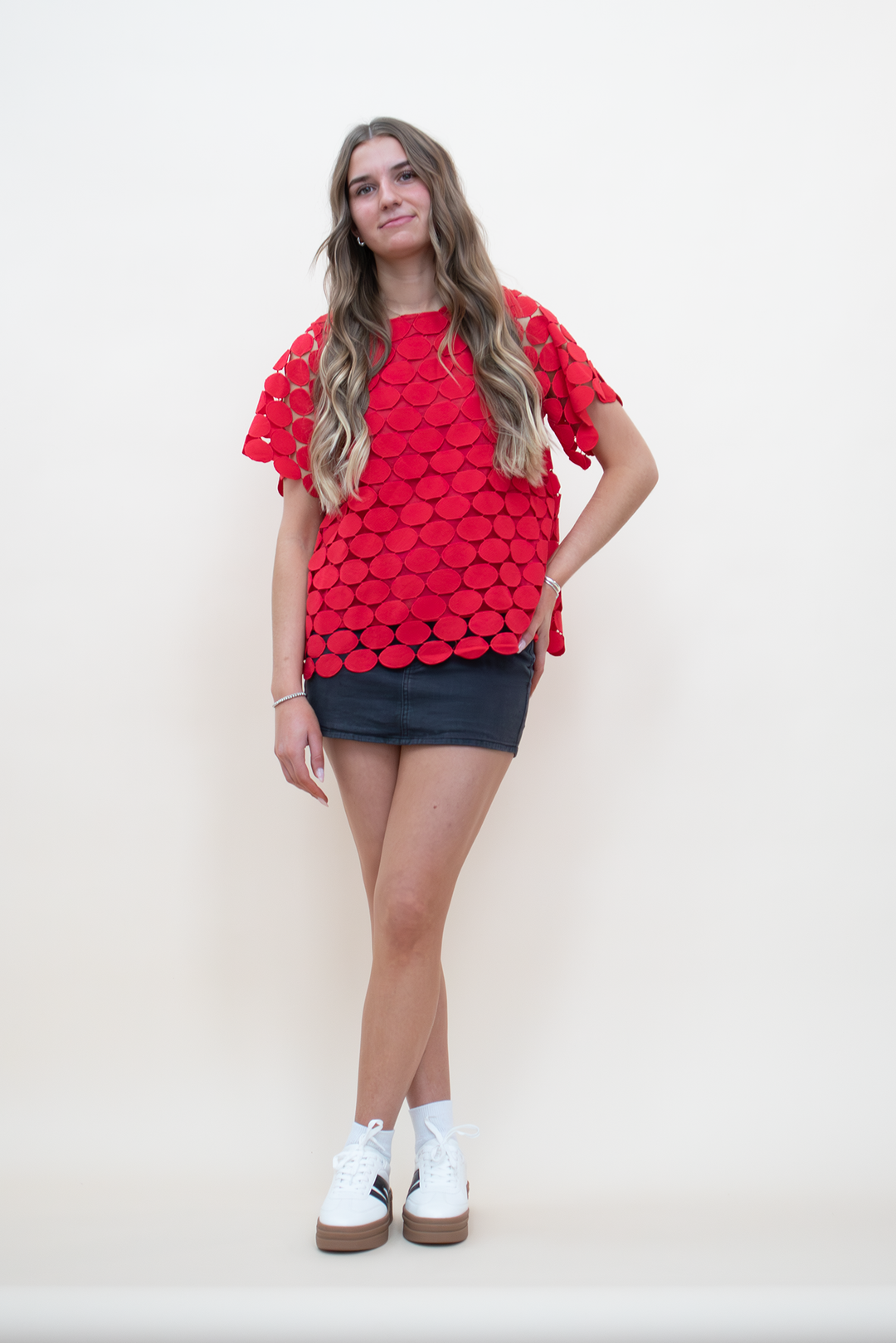 Full-length front view of red textured appliqué short-sleeve top with scalloped drop hem and semi-sheer cutwork detailing, worn over a charcoal mini skirt with white sneakers