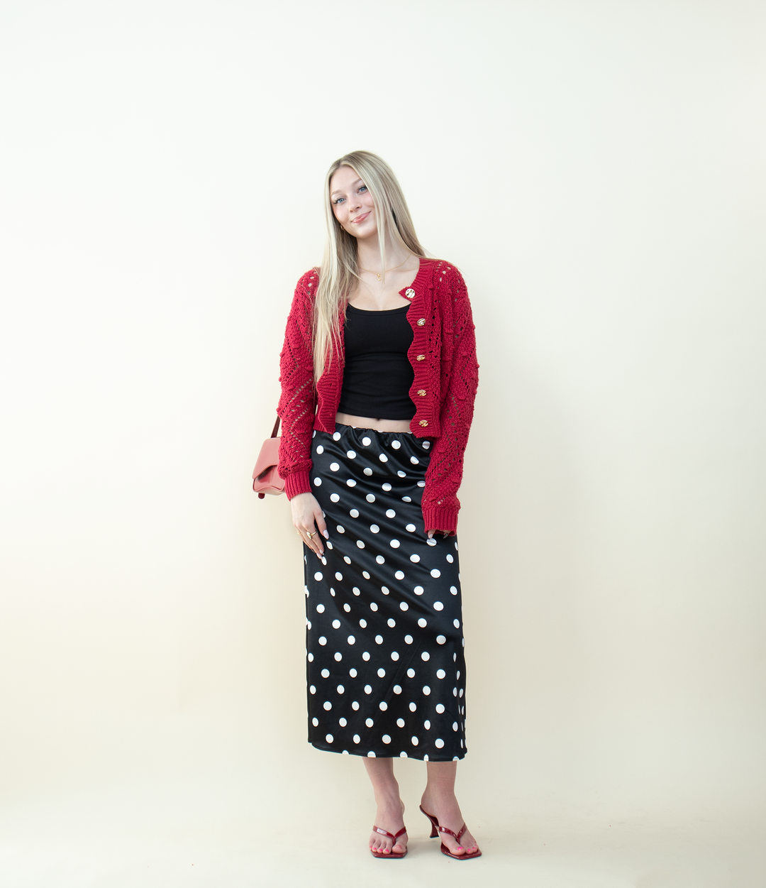 Red scalloped-edge knit cardigan worn open over a black crop top with a black-and-white polka dot skirt. Trendy layered outfit showcasing a textured red cardigan perfect for fall and winter fashion.
