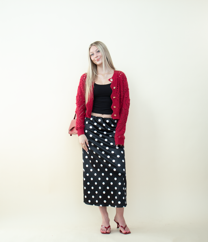 Red scalloped-edge knit cardigan worn open over a black crop top with a black-and-white polka dot skirt. Trendy layered outfit showcasing a textured red cardigan perfect for fall and winter fashion.