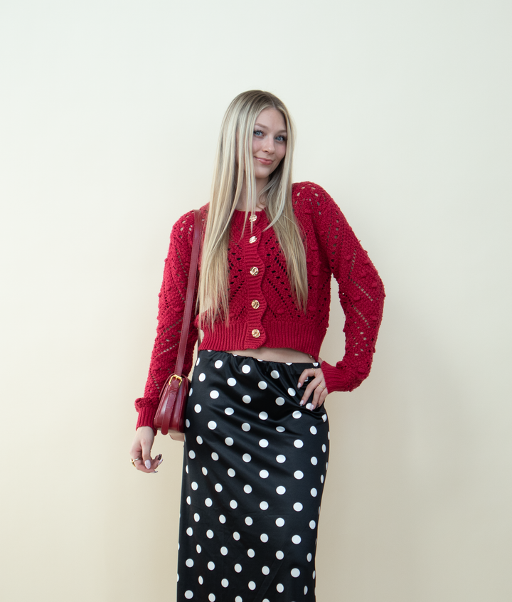 Close-up of a red chunky knit cardigan with gold buttons styled with a satin polka dot skirt and a red shoulder bag. Feminine cozy cardigan outfit idea for cold-weather style.