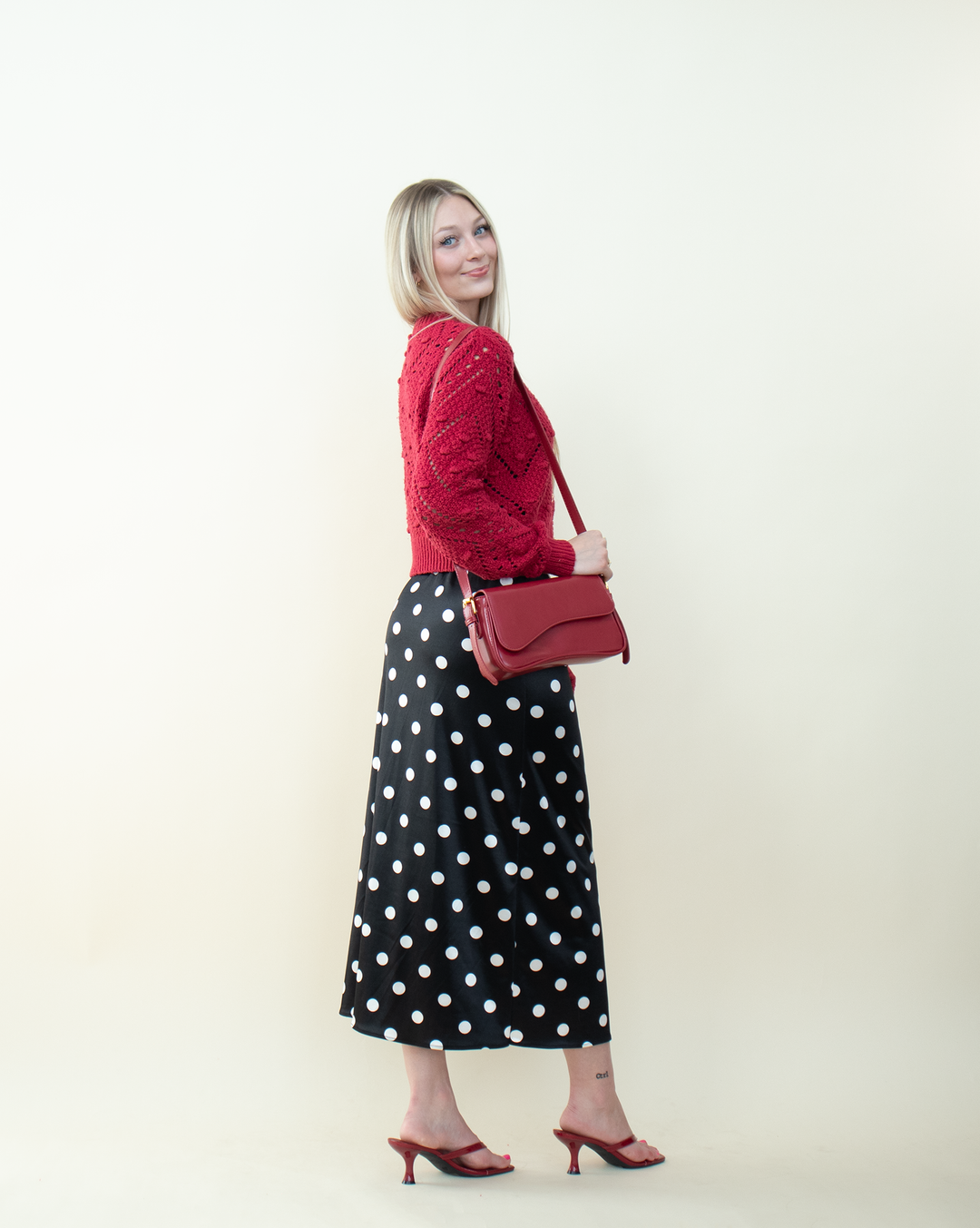 Side view of a woman wearing a bright red knit cardigan with a textured bobble-stitch design paired with a black polka dot midi skirt and red kitten-heel sandals. Modern and playful styling for a statement red cardigan.