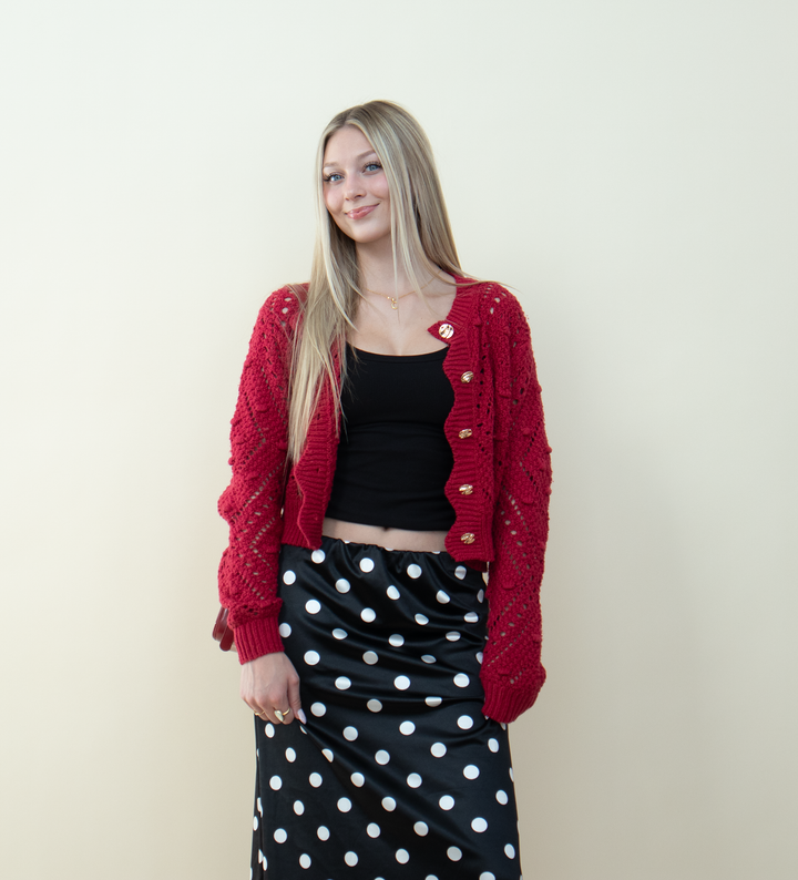 Red knitted cardigan with scalloped edges and gold buttons layered over a black tank top, styled with a black polka dot skirt. Cute and trendy red cardigan outfit inspiration for everyday wear.