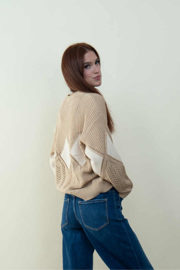 Woman with long auburn hair looking over her shoulder, wearing a beige knit sweater with white diamond patterns on the back and sleeves, standing against a plain light background.