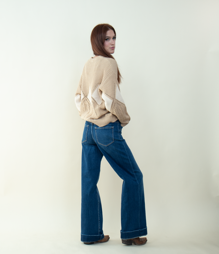 Woman posing with her back slightly turned, looking over her shoulder, wearing a beige knit sweater with white diamond patterns and blue wide-leg jeans, against a plain light background.