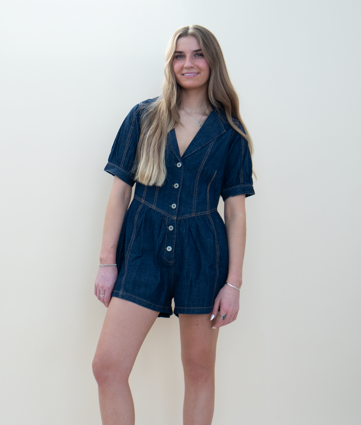 Women's navy dark wash denim romper with puff sleeves, a V-neckline with collar, button front closure, contrast topstitching, and a flared gathered short styled in front of a beige background.