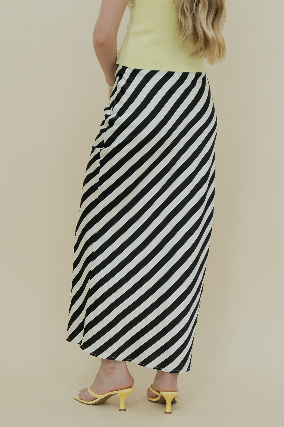 Close-up from the back view of black and white diagonal striped satin midi skirt paired with yellow heeled sandals—chic, eye-catching details perfect for summer fashion styling.
