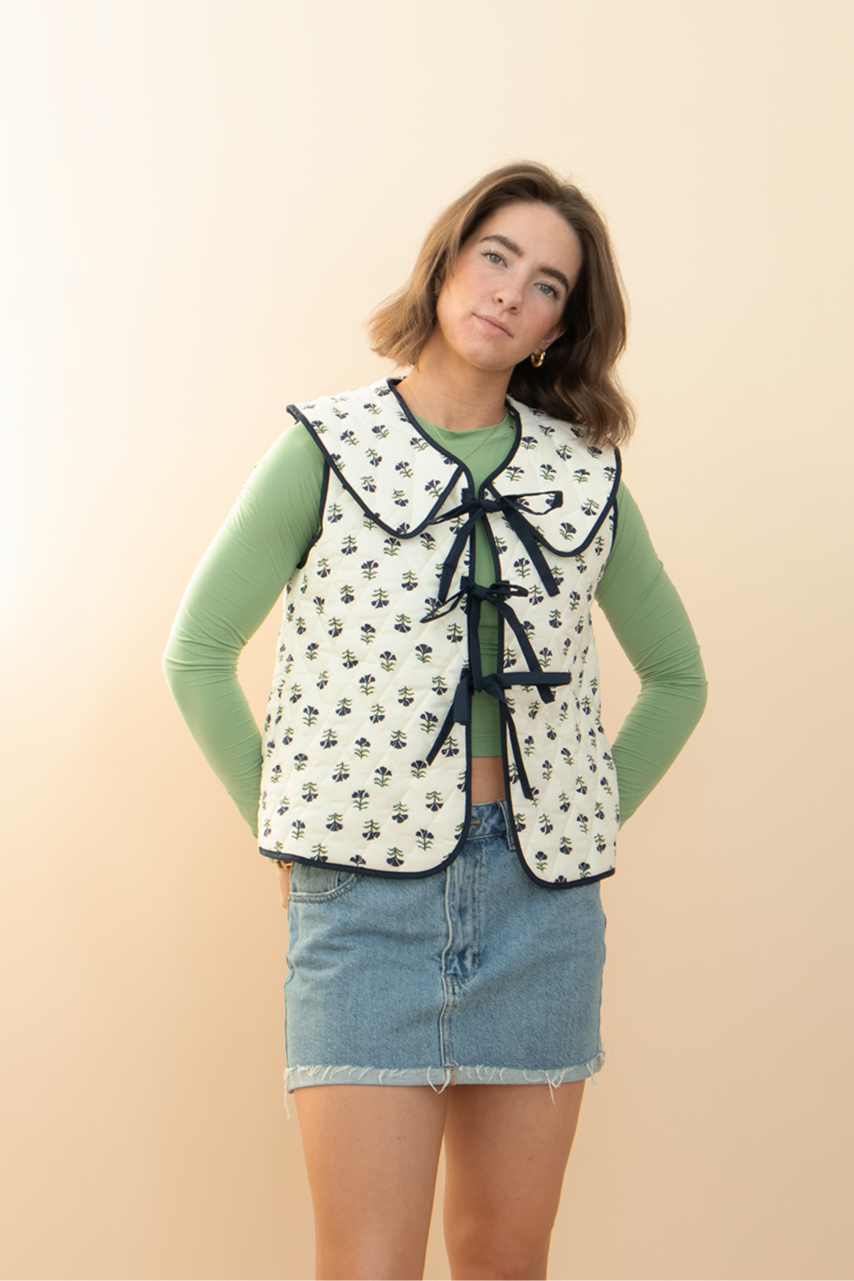 Woman wearing a floral quilted vest with black bow ties over a light green long sleeve top and denim mini skirt — modern spring outfit from Groove Closet.