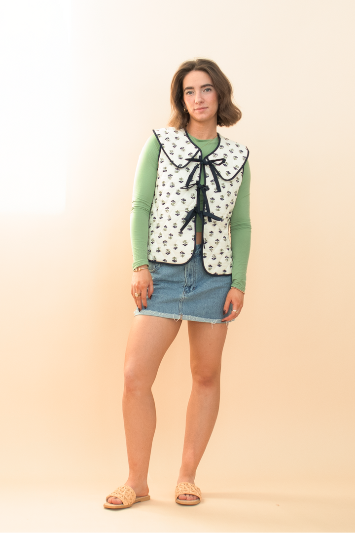 Model styled in a cottagecore-inspired outfit featuring a white floral quilted vest with navy trim, green fitted top, raw hem denim skirt, and straw slip-on heels – modern feminine fashion look.
