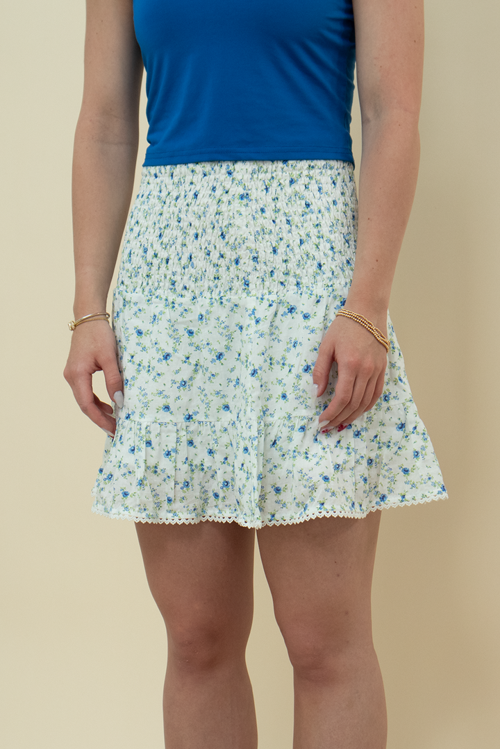 Close-up of a white mini skirt with blue floral print, smocked waistband, tiered ruffles, and lace hem detail – feminine summer skirt for women’s fashion.