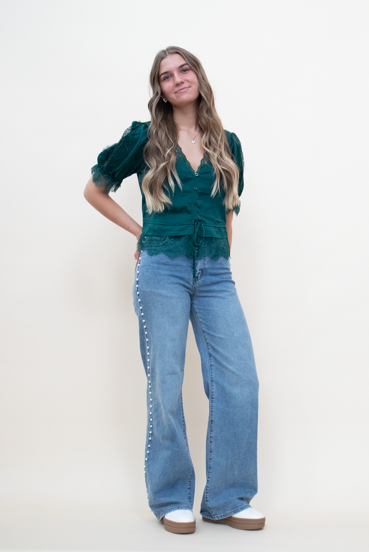 Light wash wide-leg flare jeans with pearl stud side seam trim, styled with a forest green lace puff-sleeve blouse and white platform clogs, full front view on model
