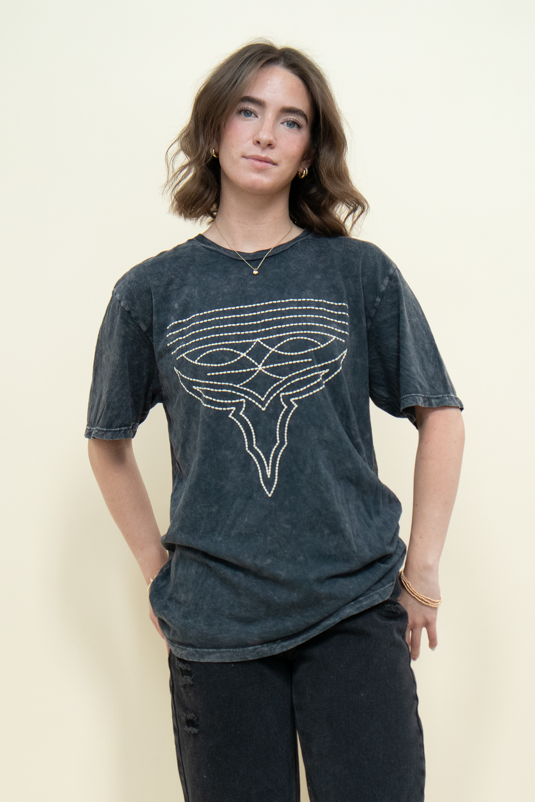 A woman modeling a dark washed graphic t-shirt with a white geometric line-art design on the front, paired with black pants, against a plain cream background.