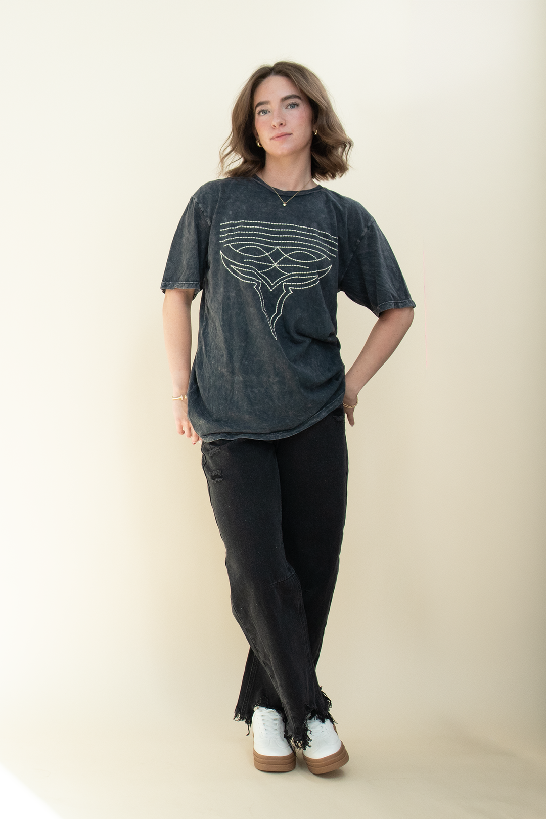 A woman modeling a dark gray mineral-wash graphic t-shirt with a white line-art design, paired with black pants and white sneakers, standing against a plain beige studio background.