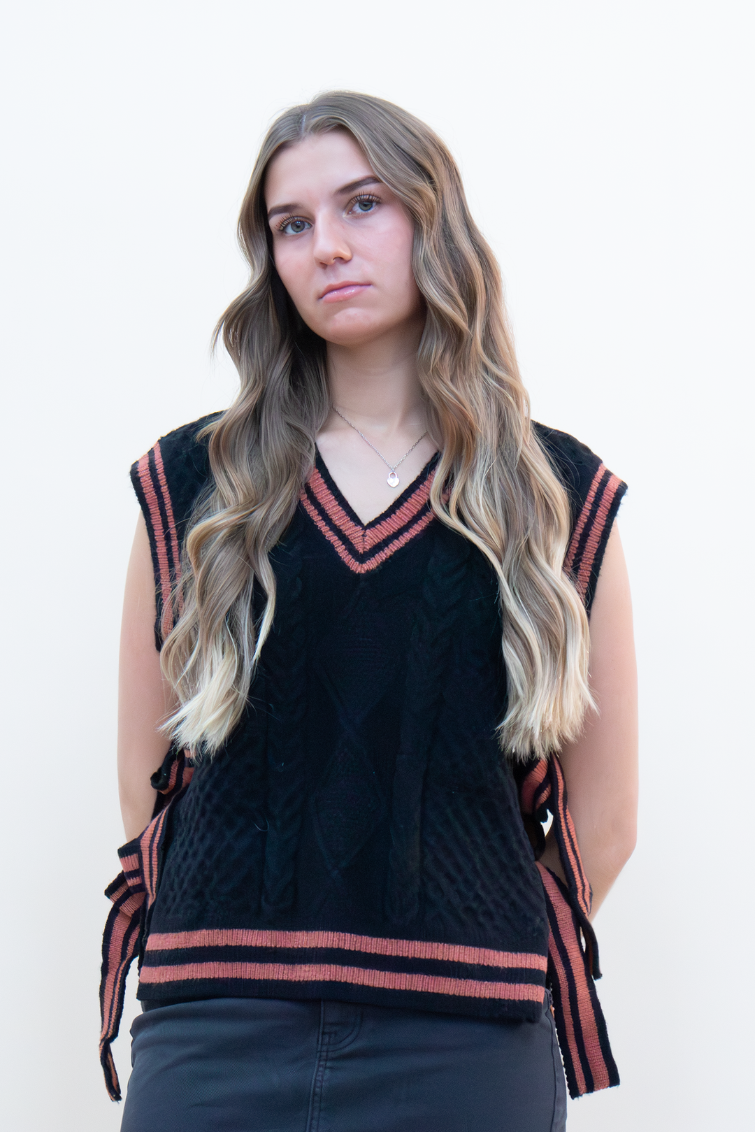 Black cable knit sweater vest with terracotta varsity stripe trim and V-neckline, styled with dark jeans on a young woman