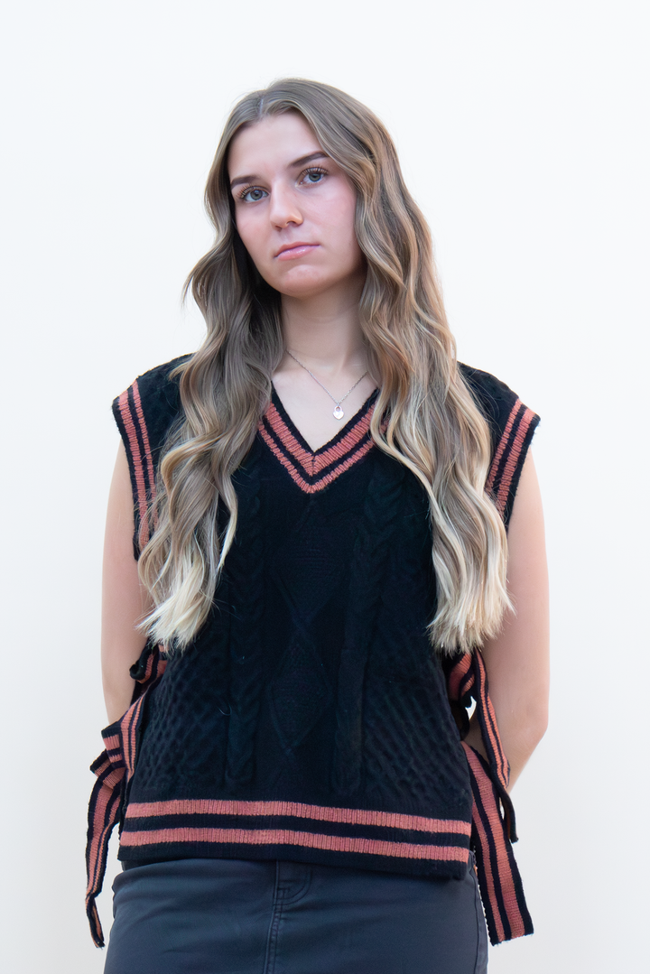 Black cable knit sweater vest with terracotta varsity stripe trim and V-neckline, styled with dark jeans on a young woman
