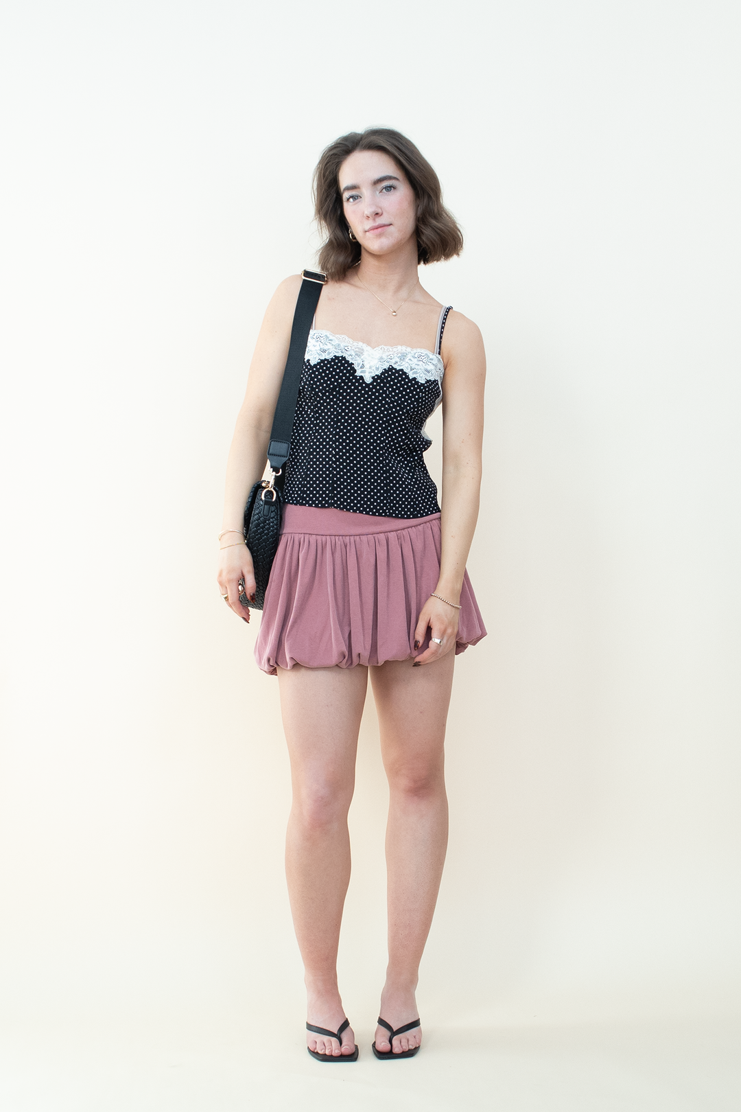 Front view of black polka dot cami with white lace neckline trim, mauve bubble mini skirt, and black kitten heel mules styled with a crossbody bag