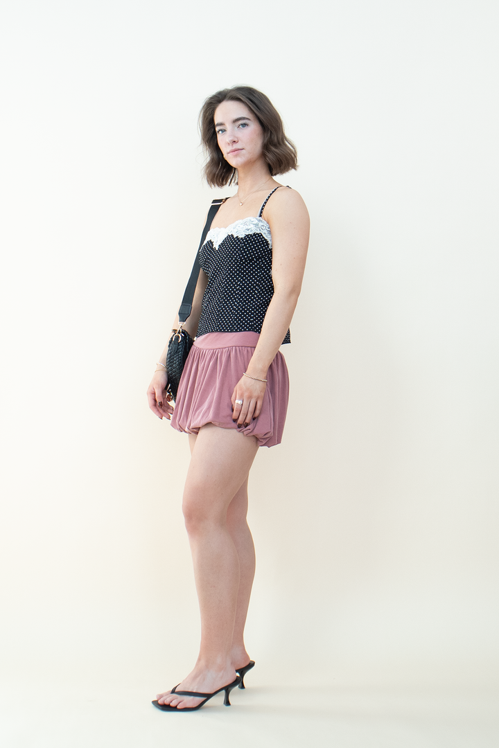 Side view of black polka dot cami top with white lace trim, styled with a mauve bubble mini skirt, black kitten heel mules, and crossbody bag