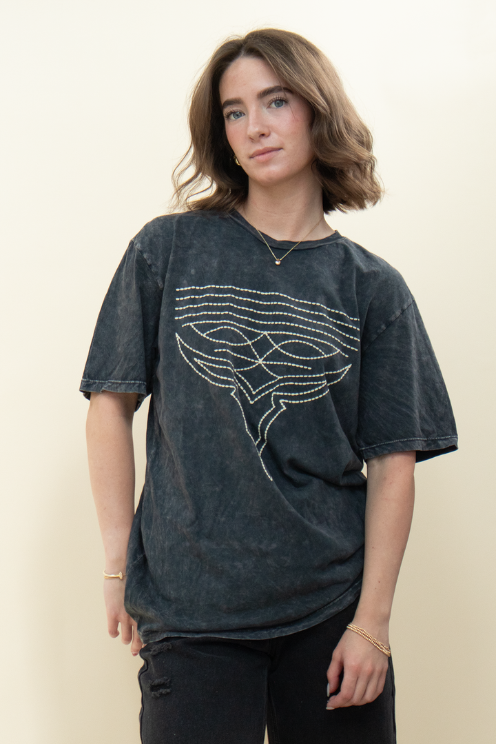 A woman modeling a dark gray mineral-wash t-shirt with a white graphic design of an oxe on the front, set against a plain beige background.