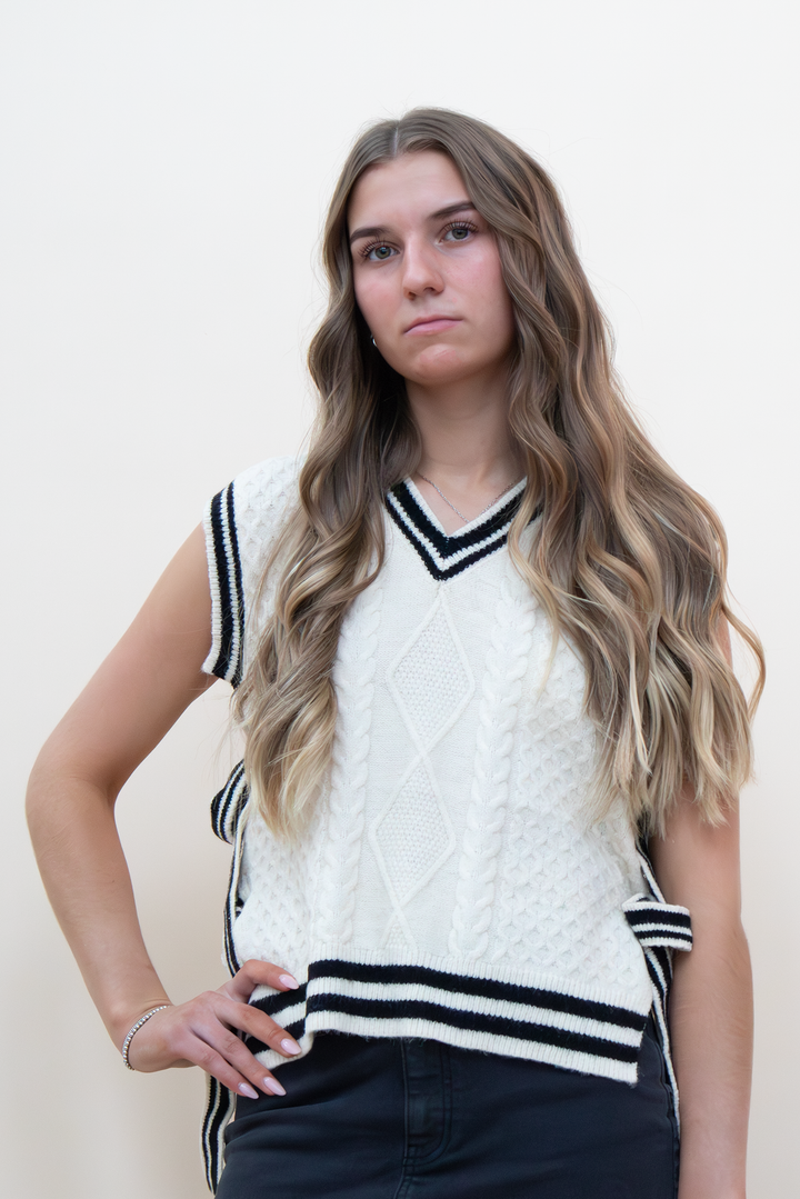 Cream cable knit sweater vest with black varsity stripe trim and V-neckline, worn by a young woman with dark jeans