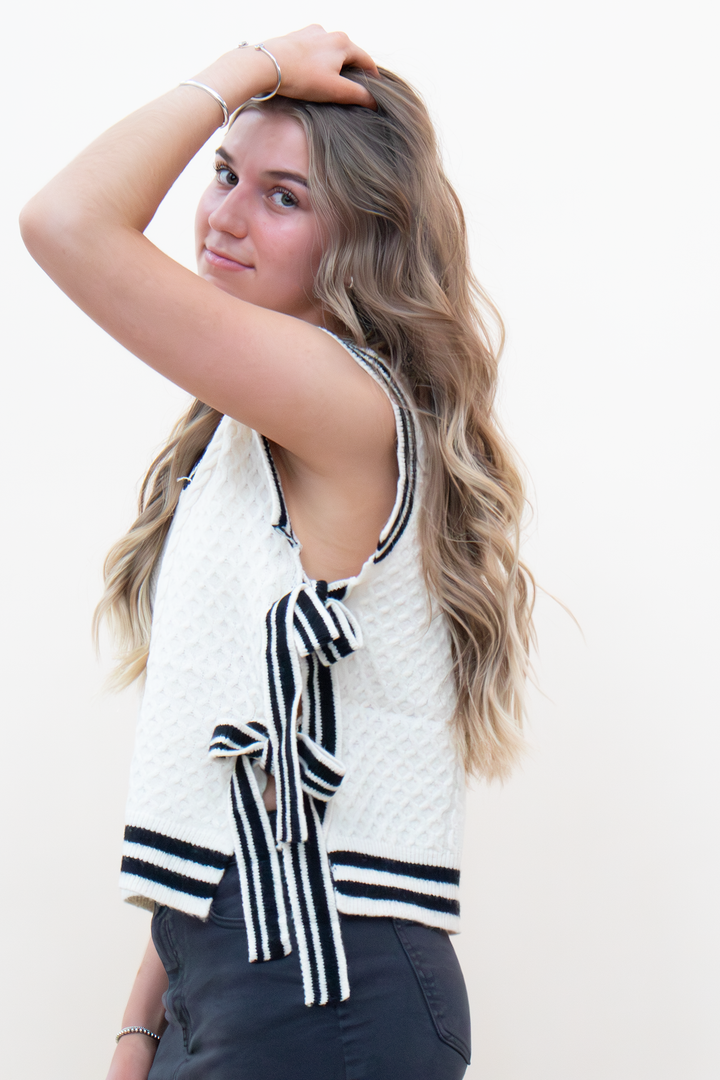 Back view of cream cable knit sweater vest featuring open back with black and white striped bow tie closure, paired with dark jeans