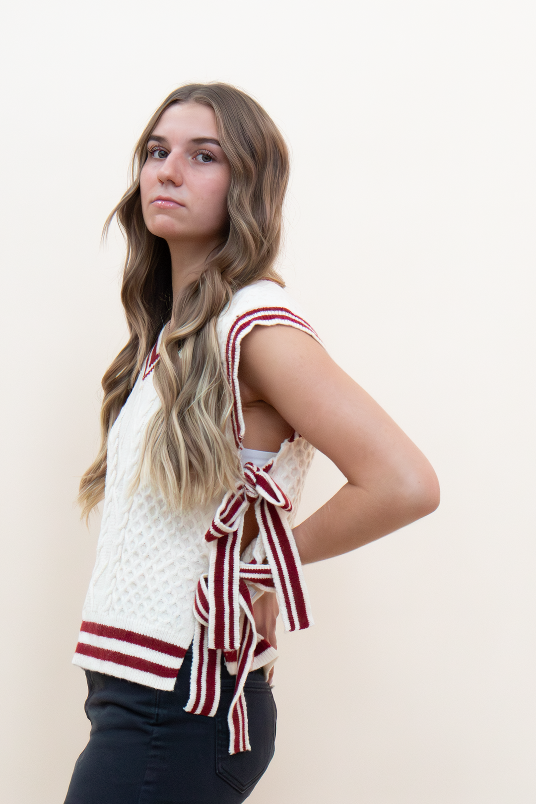 Side view of cream cable knit sweater vest with open back burgundy striped bow tie detail, worn with dark jeans