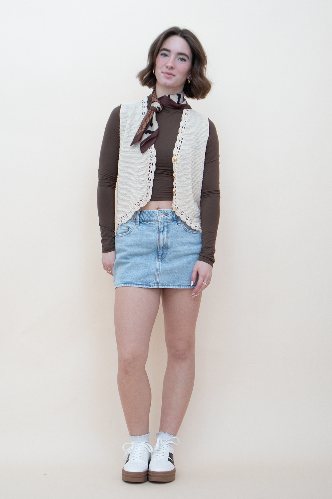 Full length view of a women's cream crochet knit button-up vest with lace trim details, worn over a brown long-sleeve top and light wash denim mini skirt