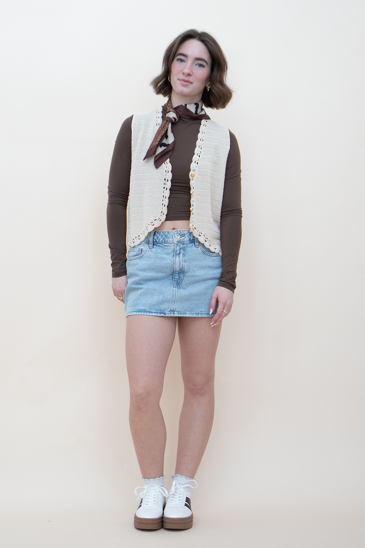 Full length view of a women's cream crochet knit button-up vest with lace trim details, worn over a brown long-sleeve top and light wash denim mini skirt